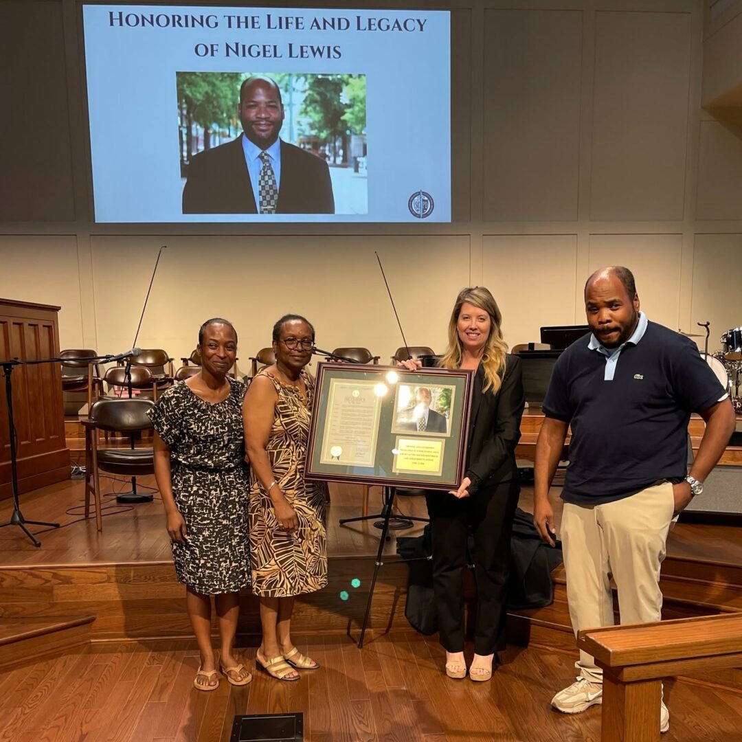 Four people stand on a church stage holding a framed tribute to Nigel Lewis, beneath a screen reading Honoring the Life and Legacy of Nigel Lewis