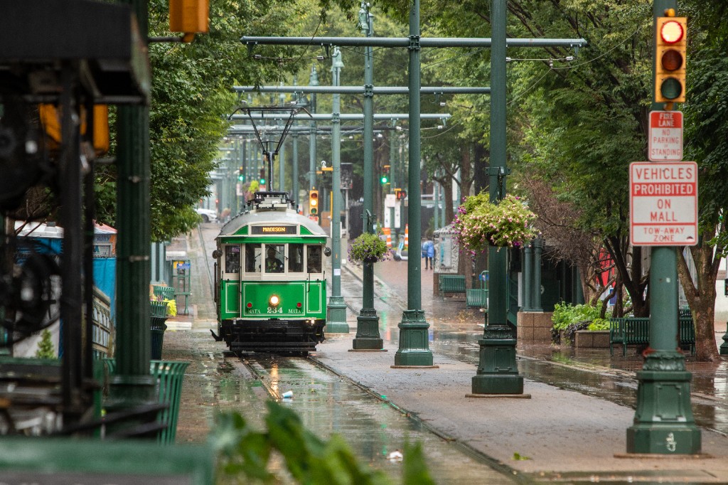 A green MATA trolley marked Madison rolls along the rain-wet Main Street Mall in downtown Memphis, lined with green lampposts and hanging flower baskets