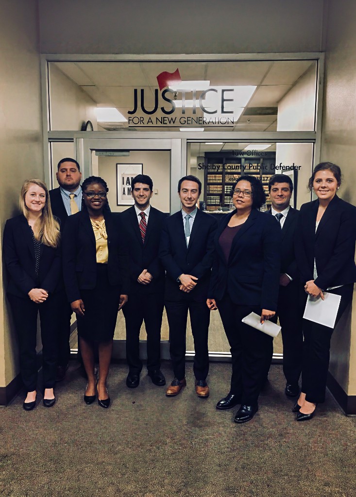 Eight members of the Shelby County Public Defender team in suits standing in the office lobby beneath a Justice for a New Generation sign
