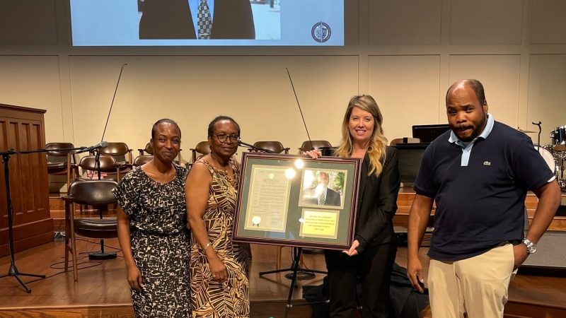 Four people stand on a church stage holding a framed tribute to Nigel Lewis, beneath a screen reading Honoring the Life and Legacy of Nigel Lewis