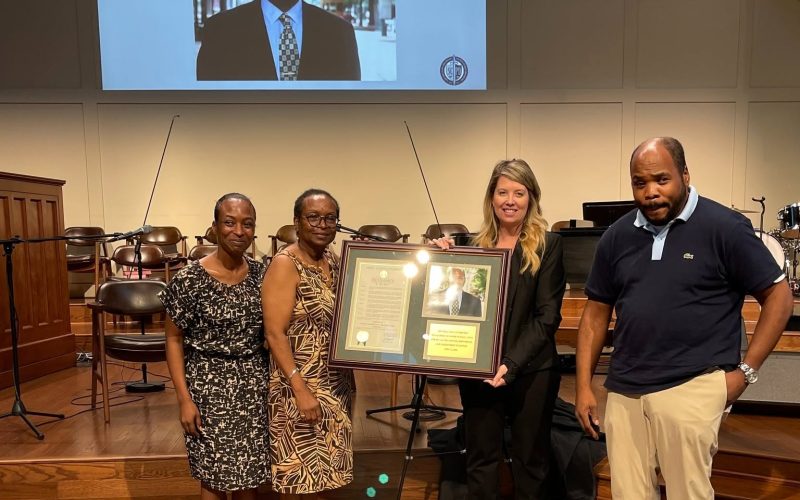 Four people stand on a church stage holding a framed tribute to Nigel Lewis, beneath a screen reading Honoring the Life and Legacy of Nigel Lewis